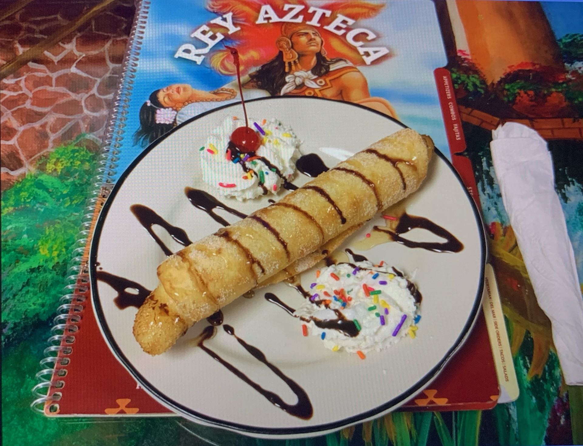 Chimi-Banana dessert featuring a deep-fried banana wrapped in a flour tortilla, topped with honey, chocolate sauce, whipped cream, and a maraschino cherry.
